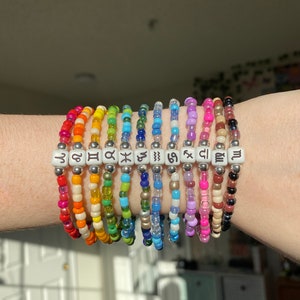 May include: A collection of beaded bracelets in a rainbow of colors. Each bracelet features a different zodiac symbol on white square beads. The bracelets are worn on a wrist.