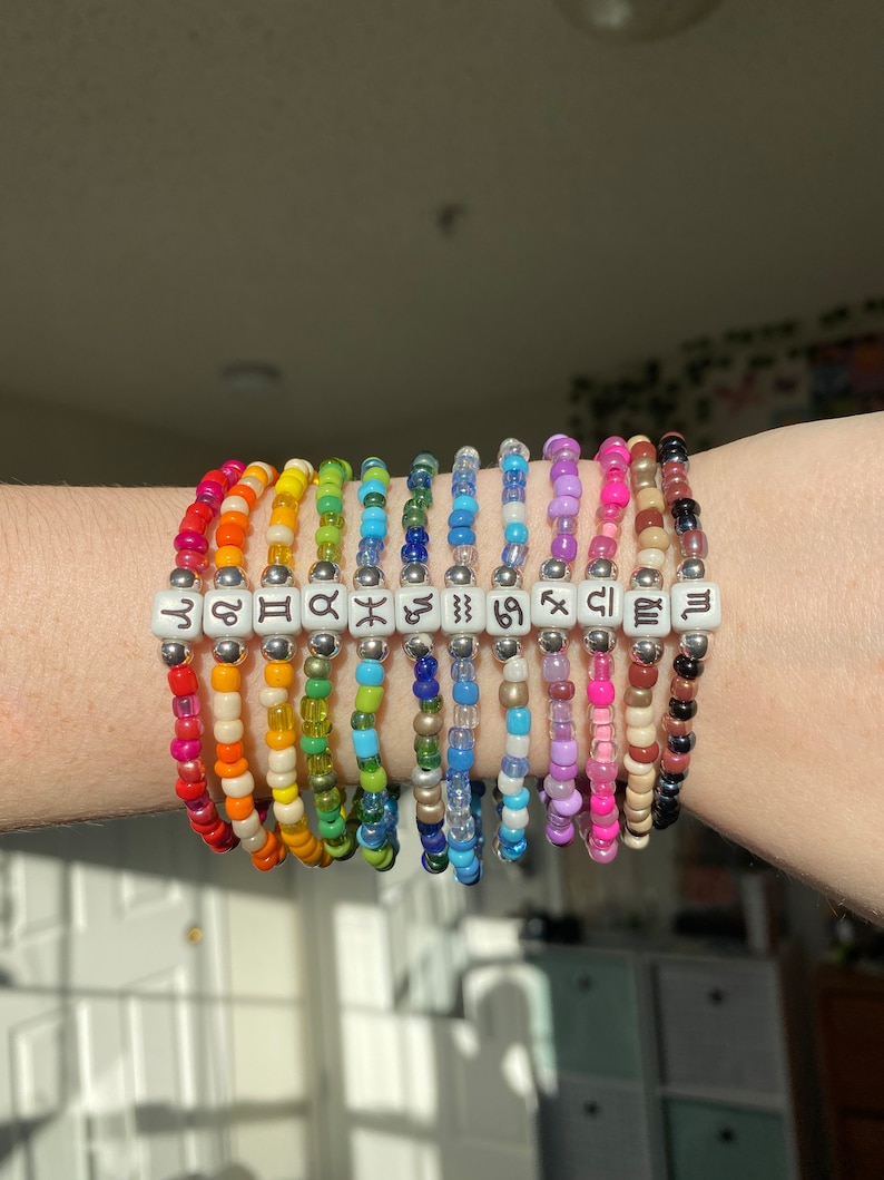 May include: A collection of beaded bracelets in a rainbow of colors. Each bracelet features a different zodiac symbol on white square beads. The bracelets are worn on a wrist.