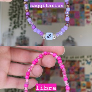 May include: Two beaded bracelets, one purple and one pink. The purple bracelet says "saggitarius" and has a Sagittarius symbol bead. The pink bracelet says "libra" and has a Libra symbol bead. Both bracelets are held up.