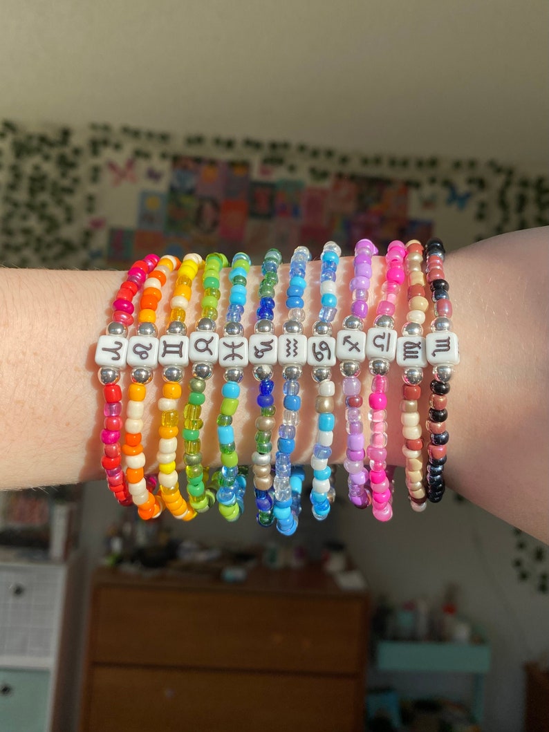 May include: A collection of beaded bracelets in a rainbow of colors. Each bracelet features a different zodiac symbol on white square beads. The bracelets are made with small, colorful beads and silver-colored spacers.