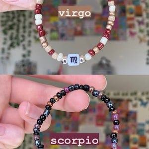 May include: Two beaded bracelets, one with burgundy, cream, and gold beads, the other with black, purple, and bronze beads. The first bracelet has a white square bead with the word "virgo" and a Virgo symbol. The second bracelet has a white square bead with the word "scorpio" and a Scorpio symbol.