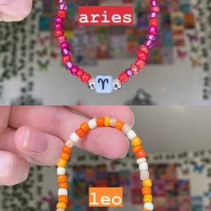 May include: Two beaded bracelets, one red and pink, the other orange and white. The top bracelet has the word "aries" and the Aries symbol. The bottom bracelet has the word "leo" and the Leo symbol. Both bracelets have a light blue bead with a silver bead on each side.