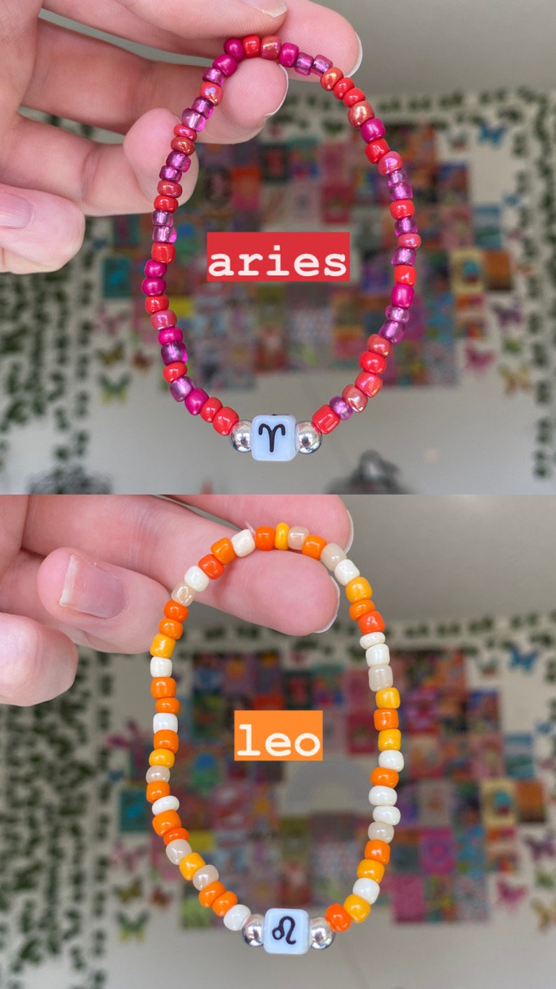 May include: Two beaded bracelets, one red and pink, the other orange and white. The top bracelet has the word "aries" and the Aries symbol. The bottom bracelet has the word "leo" and the Leo symbol. Both bracelets have a light blue bead with a silver bead on each side.