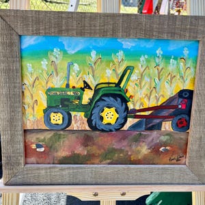 May include: A framed painting of a green John Deere 950 tractor in a field of corn. The tractor has yellow wheels and is pulling a dark plow attachment. The background features a blue sky and green corn stalks. The artist signature is in the corner.