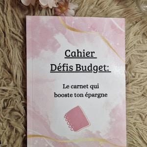 Peut inclure: Un cahier rose et blanc avec le texte "Cahier Défis Budget: Le carnet qui booste ton épargne" est présenté sur une surface texturée. Le cahier a un design abstrait rose et or et le compte Instagram "La_famille_budgetaire".