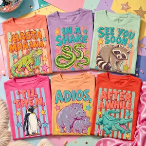 May include: Six colorful t-shirts with animal graphics and text. Designs include an iguana, snake, raccoon, penguin, hippos, and an alligator. Each shirt has a unique phrase like "Hasta Mañana" and "See You Soon."