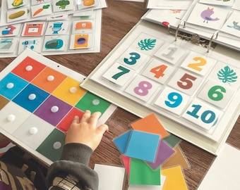 May include: A child's hands are shown placing colorful foam circles on a board with a grid of squares. The board has a variety of colors, including red, orange, yellow, green, blue, and purple. The child is also shown working with a set of number tiles, which are arranged in a grid pattern. The tiles are numbered 1 through 10. The tiles are white with black numbers. The child is also shown working with a set of colorful squares. The squares are arranged in a random pattern. The squares are blue, green, yellow, and orange.