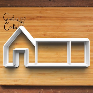 Puede incluir: Cortador de galletas de plástico blanco con forma de casa con una ventana y una puerta. El cortador de galletas está sobre una superficie de madera.