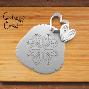 May include: A white plastic cookie cutter set on a wooden surface. The set includes a large, irregularly shaped cutter with a floral design and a heart-shaped cutter. The text "Cactus Cookies" is in the upper left corner.