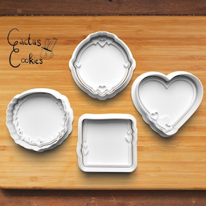 May include: Four white plastic cookie cutters on a wooden surface. The cutters are shaped like a heart, a square, and two different round frames with heart details.