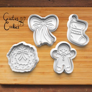 Op de afbeelding: Vier witte plastic kerstkoekjesvormen op een houten oppervlak. De vormen zijn in de vorm van een peperkoekman, een sok, een strik en een krans.