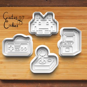Może przedstawiać: Cztery białe plastikowe wykrawacze do ciastek w kształcie kontrolerów do gier retro i automatów do gier. Wykrawacze są ułożone na drewnianej desce do krojenia. Tekst "Cactus Cookies" znajduje się w lewym górnym rogu.