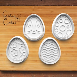 Peut inclure: Quatre emportes-pièces en plastique blanc en forme d'œuf de Pâques sur une surface en bois. Les emportes-pièces présentent différents motifs : un visage de lapin, un motif de lignes ondulées, un motif floral et un motif circulaire.