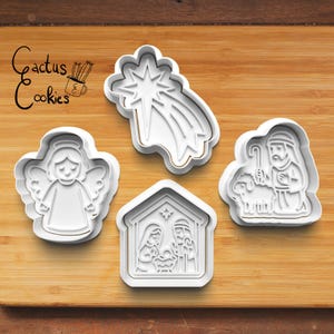 May include: Four white plastic Christmas cookie cutters on a wooden surface. The cutters include an angel, a star with a tail, a nativity scene, and a shepherd with a sheep. The text "Cactus Cookies" is in the upper left corner.