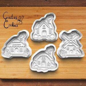 May include: Four white plastic cookie cutters in the shapes of houses and a windmill, arranged on a wooden surface. The cutters have detailed designs of windows, doors, and chimneys. The text "Cactus Cookies" is in the upper left corner.