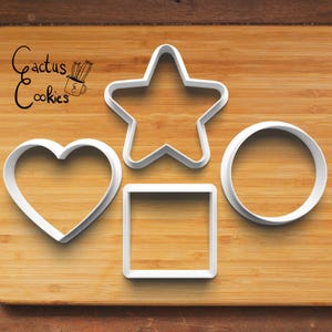 Może przedstawiać: Cztery białe plastikowe wykrawacze do ciastek w kształcie serca, gwiazdy, kwadratu i koła ułożone na drewnianej powierzchni. Tekst "Cactus Cookies" jest widoczny w lewym górnym rogu.