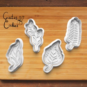 Puede incluir: Cuatro cortadores de galletas de plástico blanco con forma de hojas sobre una superficie de madera. Los cortadores son para hacer galletas con forma de hojas. El texto "Cactus Cookies" es visible en la esquina superior izquierda de la imagen.