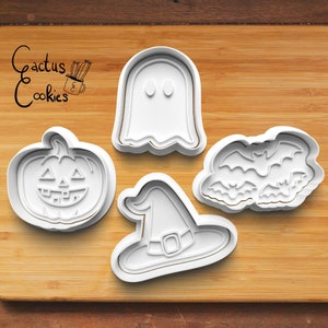 Puede incluir: Cuatro cortadores de galletas blancos con temática de Halloween sobre una superficie de madera. Las formas incluyen una calabaza, un fantasma, un sombrero de bruja y un grupo de murciélagos. La marca "Cactus Cookies" está escrita en cursiva.