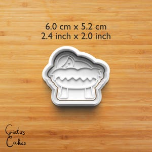 Puede incluir: Un cortador de galletas de pl&aacute;stico blanco con forma de escena de la Natividad, que mide 6,0 cm por 5,2 cm. El dise&ntilde;o incluye un beb&eacute; en un pesebre. El cortador est&aacute; sobre una superficie de madera. El texto "Cactus Cookies" est&aacute; en la esquina inferior izquierda.