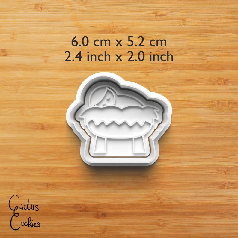 Puede incluir: Un cortador de galletas de pl&aacute;stico blanco con forma de escena de la Natividad, que mide 6,0 cm por 5,2 cm. El dise&ntilde;o incluye un beb&eacute; en un pesebre. El cortador est&aacute; sobre una superficie de madera. El texto "Cactus Cookies" est&aacute; en la esquina inferior izquierda.