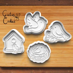 May include: Four white plastic cookie cutters in bird-themed shapes: a bird in flight, a bird on a branch, a bird in a nest, and a birdhouse with a bird. The logo "Cactus Cookies" is in the upper left corner.