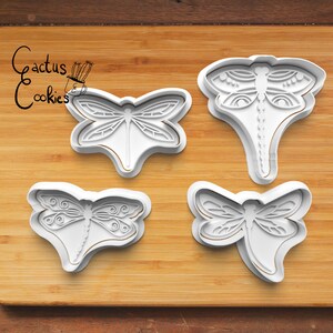 May include: Four white plastic dragonfly cookie cutters on a wooden cutting board. The cookie cutters are different sizes and have intricate designs.