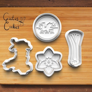 May include: Four white plastic cookie cutters on a wooden surface. The cutters are shaped like a dragon, a daffodil, a leek, and the outline of Wales.