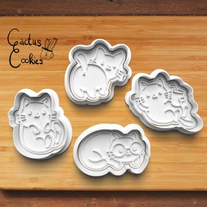 Puede incluir: Cuatro cortadores de galletas blancos con forma de gato sobre una superficie de madera. Los cortadores están en diferentes poses, incluyendo un gato acurrucado, un gato con una pata levantada y un gato acostado. El texto "Cactus Cookies" está en la esquina superior izquierda.