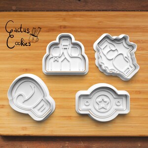 May include: Four white plastic cookie cutters on a wooden surface. The cutters are shaped like a boxing glove, a boxing belt, a group of three people raising a bottle, and two people raising their fists.