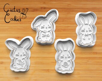 May include: Four white plastic bunny cookie cutters on a wooden surface. The cutters feature different bunny designs, including one holding a flower, one holding a carrot, one holding an egg, and one with a bow tie.