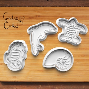 Op de afbeelding: Vier witte plastic koekjesvormen in de vorm van een zeepaardje, een dolfijn, een schildpad en een nautilus schelp. De vormen liggen op een houten snijplank met de tekst "Cactus Cookies" in zwarte inkt.