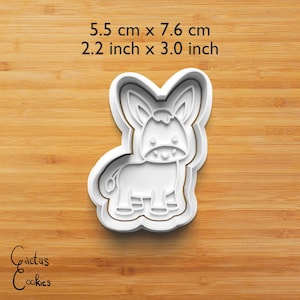 Puede incluir: Cortador de galletas blanco con forma de burro, de 5,5 cm por 7,6 cm (2,2 pulgadas x 3,0 pulgadas). El dise&ntilde;o presenta un burro sonriente con orejas grandes y una cola peque&ntilde;a. El texto "Cactus Cookies" est&aacute; en la esquina inferior izquierda.