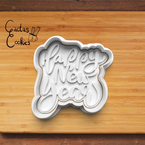 May include: White plastic cookie cutter in the shape of "Happy New Year" in a cursive font. The cutter has a layered design and is set against a wooden background. The Cactus Cookies logo is in the upper left corner.