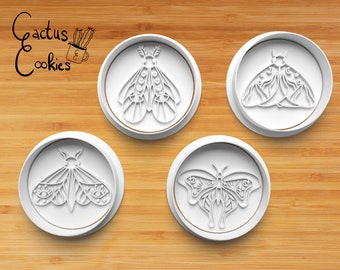 May include: Four white plastic cookie cutters on a wooden surface. The cutters are shaped like moths and butterflies with intricate designs. The text "Cactus Cookies" is visible in the top left corner of the image.