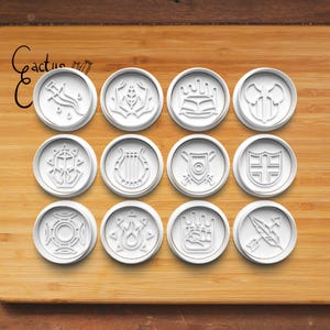 May include: Twelve white round cookie cutters with various designs, including a crown, a lyre, a sword, and a hand. The cookie cutters are arranged in three rows of four on a wooden surface.