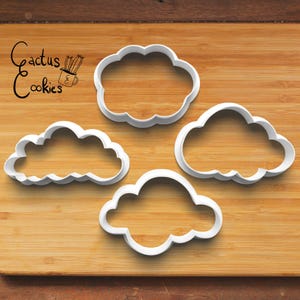May include: Four white plastic cloud-shaped cookie cutters on a wooden surface. The cookie cutters are different sizes and shapes.