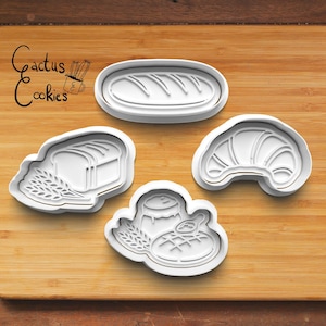 May include: Four white plastic cookie cutters in the shapes of bread, a loaf of bread, a croissant, and a dessert with a spoon. The Cactus Cookies logo is in the upper left corner. The cutters are on a wooden surface.