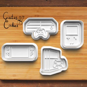 Może przedstawiać: Cztery białe plastikowe foremki do ciasteczek w kształcie konsol do gier wideo i kontrolerów. Formy znajdują się na drewnianej powierzchni.