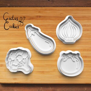 May include: Four white plastic cookie cutters shaped like vegetables: an eggplant, an onion, a tomato, and a broccoli. The cutters are on a wooden cutting board with the text "Cactus Cookies" in black ink.