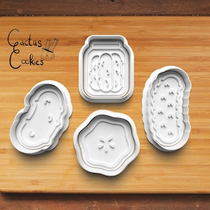 May include: Four white plastic cookie cutters shaped like a jar of pickles, a pickle, a pickle slice, and a pickle spear. The cutters are on a wooden surface.