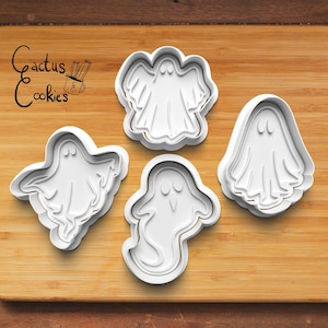 Op de afbeelding: Vier witte plastic koekjesvormen in de vorm van een spook op een houten snijplank. De koekjesvormen zijn in verschillende maten en vormen. De tekst "Cactus Cookies" staat op de plank.