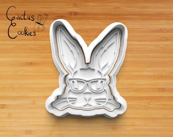Peut inclure: Emporte-pièce en plastique blanc en forme de lapin portant des lunettes de soleil. Le lapin a de grandes oreilles, un petit nez et un large sourire.