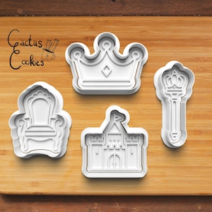 May include: Four white plastic cookie cutters in the shapes of a crown, a throne, a castle, and a scepter. The cookie cutters are arranged on a wooden surface. The text "Cactus Cookies" is in the upper left corner.