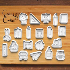 Puede incluir: Un conjunto de cortadores de galletas de plástico blanco con formas escolares, como un globo terráqueo, una regla, un lápiz, una manzana y un autobús escolar. El texto "Cactus Cookies" está escrito en cursiva. Los cortadores están dispuestos sobre una superficie de madera.