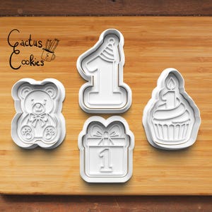 May include: Four white plastic cookie cutters in the shapes of a teddy bear, the number one with a party hat, a gift box with the number one, and a cupcake with a candle. The Cactus Cookies logo is in the upper left corner.