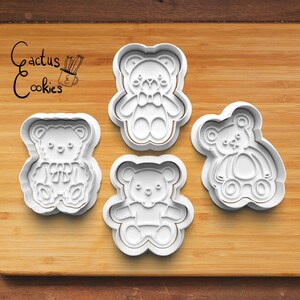 May include: Four white plastic cookie cutters shaped like teddy bears. The cutters are on a wooden surface. The text "Cactus Cookies" is written in black on the surface.
