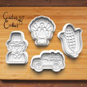 May include: Set of four white plastic cookie cutters shaped like a pilgrim, a turkey, a corn cob, and a pickup truck carrying pumpkins. The cutters are on a wooden surface.