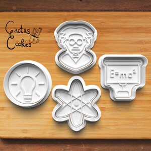 May include: Four white plastic cookie cutters in science-themed shapes: a lightbulb, an atom, a scientist, and a chalkboard with the equation E=mc². The Cactus Cookies logo is in the upper left corner.
