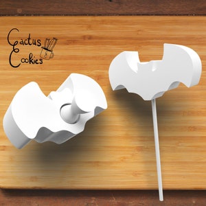 May include: Two white plastic bat-shaped cookie cutters on a wooden surface. One cutter is on a stick, the other is laying flat. The cutters are for making Halloween cookies.
