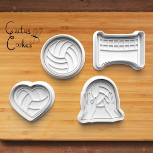 May include: Four white plastic cookie cutters shaped like a volleyball, a volleyball in a heart, a volleyball net, and a person holding a volleyball. The cutters are on a wooden surface.
